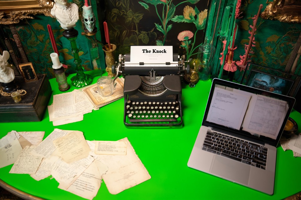 A typewriter and scattered manuscript pages: the novel a session of The Knock produces.